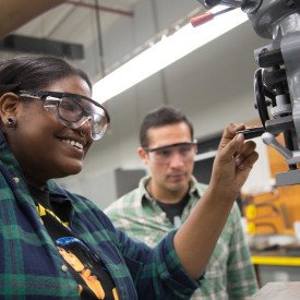 Student smiling while operating a machine with another student paying close attention over her shoulder